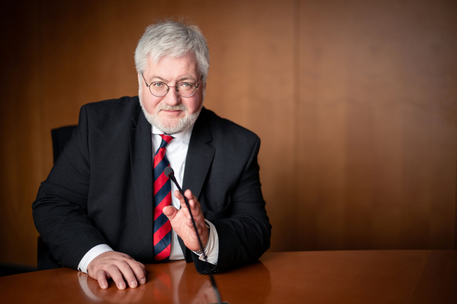 Stephan Albani, trägt Brille, schwarzen Anzug und eine rot-blau gestreifte Krawatte. Er sitzt an einem Konferenztisch, blickt freundlich in die Kamera und gestikuliert mit einer Hand in Richtung eines Mikrofons. Der Hintergrund ist aus warmem Holz gestaltet.